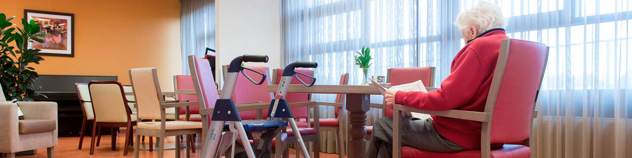 Een senior bewoner zit in de huiskamer van een verpleeghuis met naast haar een rollator